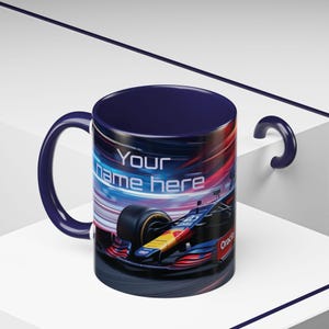 Puede incluir: Una taza de cerámica azul con un diseño de coche de carreras. El coche es rojo, amarillo y azul con el texto "Oracle" en el lateral. La taza tiene el texto "Your name here" en el lateral.