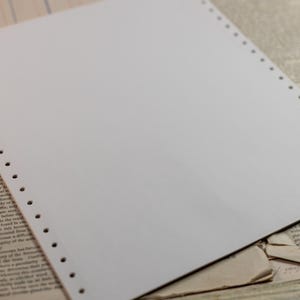 May include: A single sheet of blank white computer paper featuring a vertical row of perforated tractor-feed holes along the left edge. The paper is positioned at an angle, displaying a clean, smooth surface intended for dot matrix printing.