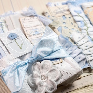 Könnte beinhalten: Eine Sammlung geschichteter Stoffreste und Papier-Ephemera in hellblauen und weißen Tönen. Die Teile umfassen Spitzenbesatz, Ösenstoff, eine hellblaue Schleife mit Punkten, einen blumenförmigen Knopf und Karten mit dem Text Swatches.