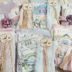 May include: A collection of fabric bundles featuring house-shaped paper cutouts, lace trim, and fabric flowers. Each bundle includes a small tag with a bird illustration and the text 25. The textiles are in shades of blue, pink, and white.