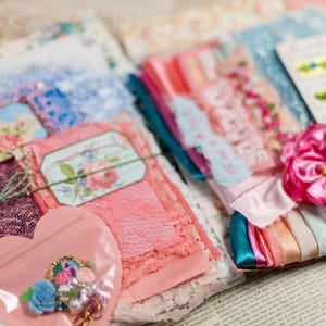May include: A collection of craft supplies featuring layered pink and blue lace, fabric scraps, and sequined material. A heart-shaped plastic pouch contains small floral embellishments and beads. A large pink fabric flower sits atop ribbons.