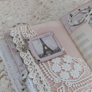 May include: A handmade journal with a vintage aesthetic. The open pages reveal a soft pink interior, adorned with lace, a small Eiffel Tower image, and delicate embellishments. A ribbon and charm add a touch of elegance to this unique keepsake.