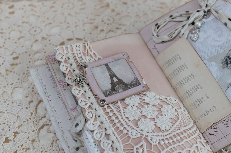May include: A handmade journal with a vintage aesthetic. The open pages reveal a soft pink interior, adorned with lace, a small Eiffel Tower image, and delicate embellishments. A ribbon and charm add a touch of elegance to this unique keepsake.