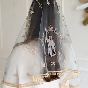 May include: A sheer, light blue veil with gold floral embroidery and a scalloped edge. The veil features a detailed embroidered image of a saint holding a sword. The design includes small flower and star patterns.