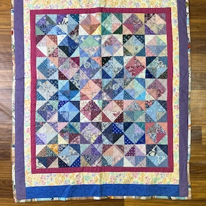 Pode incluir: Um edredom feito à mão com um design de retalhos. O edredom apresenta um quadrado central de retalhos em forma de diamante em vários tons de azul, roxo, rosa e verde. É emoldurado por um tecido rosa, floral e azul.