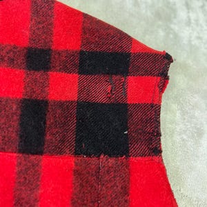 May include: A red and black plaid flannel shirt featuring a collar and a small visible tear in the fabric near the shoulder area. The garment is constructed from a woven textile with a distinct checkered pattern throughout the material.
