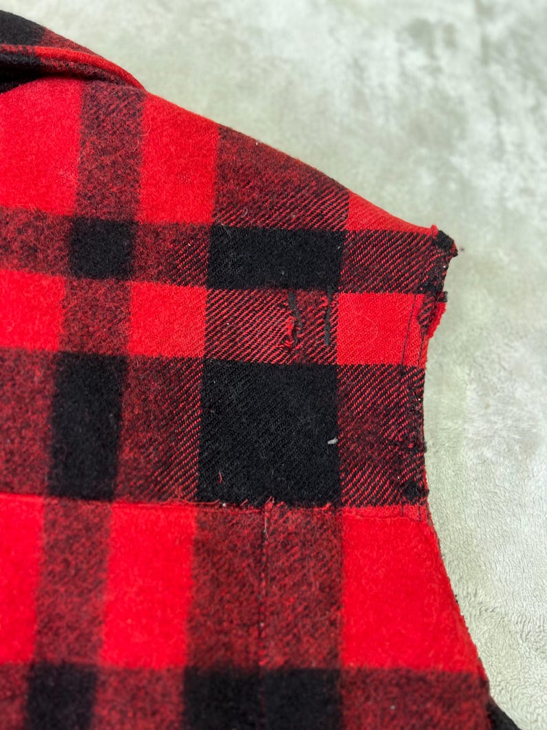 May include: A red and black plaid flannel shirt featuring a collar and a small visible tear in the fabric near the shoulder area. The garment is constructed from a woven textile with a distinct checkered pattern throughout the material.