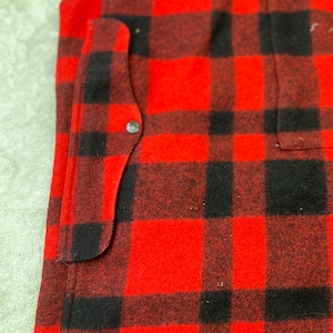 May include: A red and black buffalo plaid vest featuring a thick, textured fabric construction. The garment includes a vertical side pocket with a single silver-toned metal snap button closure, visible along the front edge of the plaid pattern.