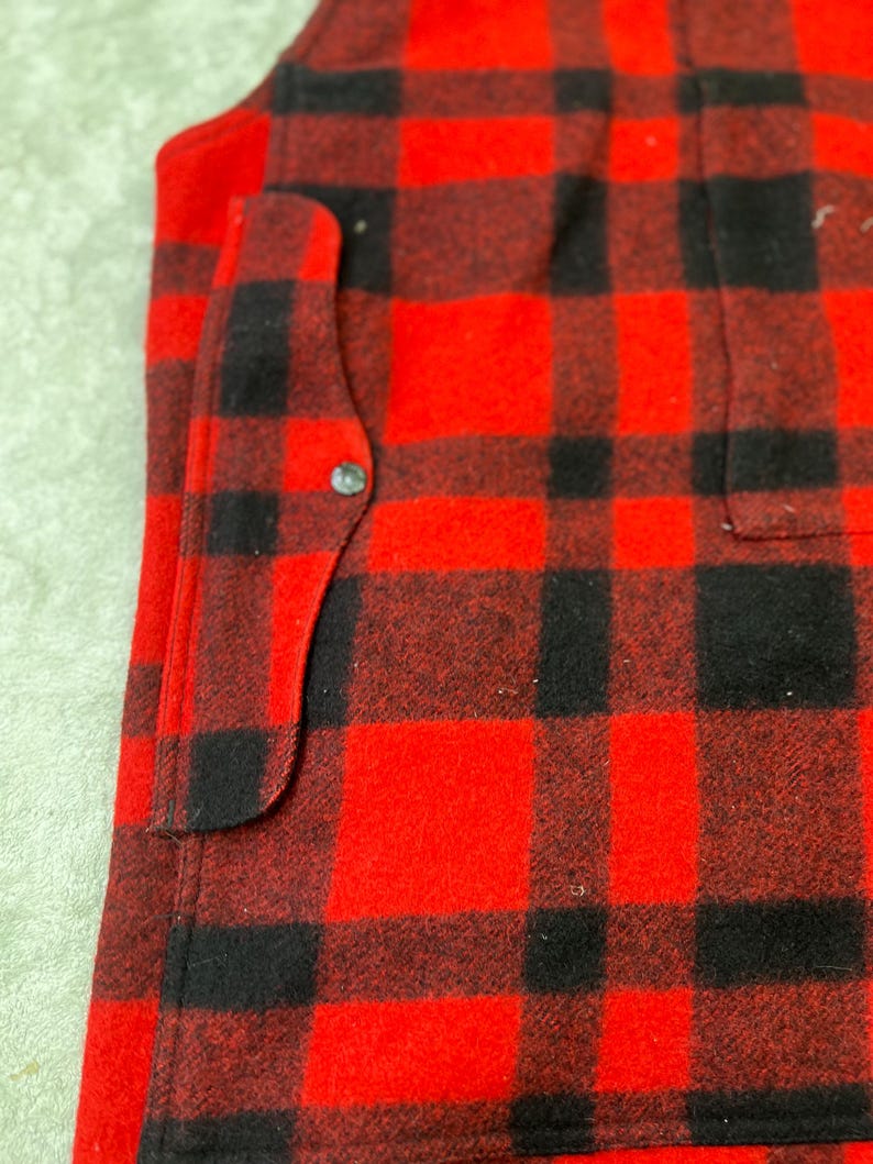 May include: A red and black buffalo plaid vest featuring a thick, textured fabric construction. The garment includes a vertical side pocket with a single silver-toned metal snap button closure, visible along the front edge of the plaid pattern.