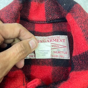 May include: A red and black buffalo plaid wool shirt featuring a white interior label that reads THIS IS A GENUINE FILSON GARMENT, SEATTLE, and a smaller tag stating Professionally Dry clean only. The shirt has a classic collar design.
