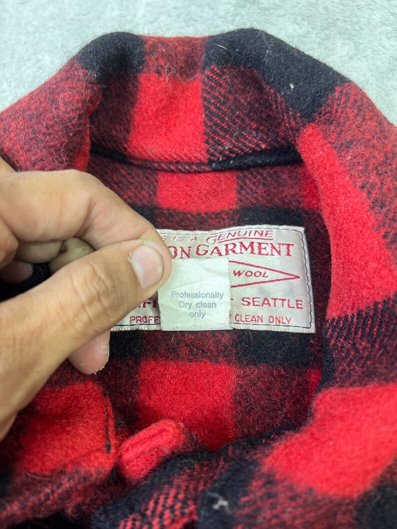 May include: A red and black buffalo plaid wool shirt featuring a white interior label that reads THIS IS A GENUINE FILSON GARMENT, SEATTLE, and a smaller tag stating Professionally Dry clean only. The shirt has a classic collar design.