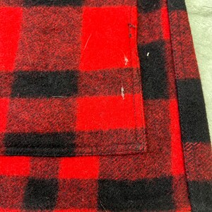 May include: A red and black buffalo plaid fabric garment featuring a thick, textured weave. The material displays a repeating square pattern with visible stitching along the vertical edge and a rectangular pocket section on the front surface.