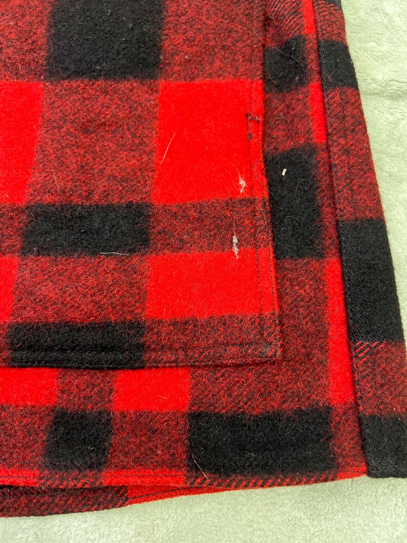 May include: A red and black buffalo plaid fabric garment featuring a thick, textured weave. The material displays a repeating square pattern with visible stitching along the vertical edge and a rectangular pocket section on the front surface.