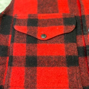 May include: A red and black buffalo plaid vest featuring a front pocket with a rounded flap and a single dark button closure. The garment is constructed from a thick, textured fabric and includes a vertical row of dark buttons along the front edge.