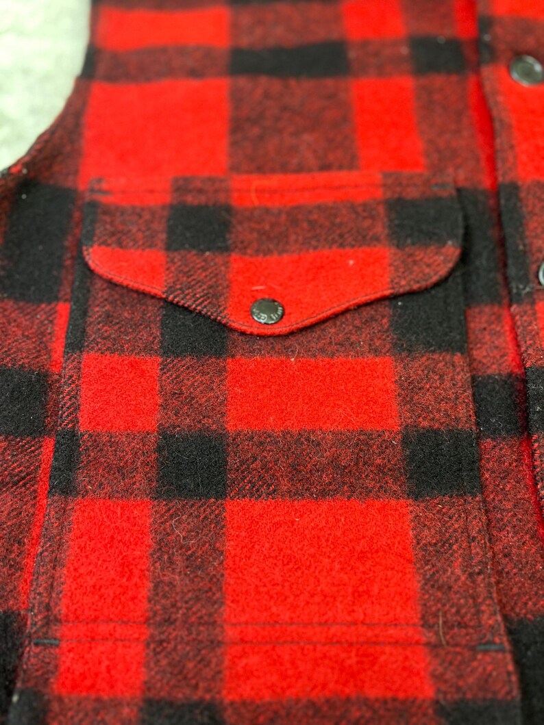 May include: A red and black buffalo plaid vest featuring a front pocket with a rounded flap and a single dark button closure. The garment is constructed from a thick, textured fabric and includes a vertical row of dark buttons along the front edge.