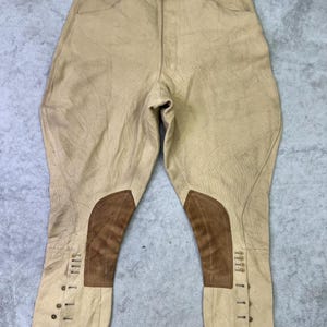 May include: Beige breeches featuring brown suede knee patches and a button-up fly. The garment includes a high waistband with multiple buttons and a series of small buttons along the lower outer leg seams for a secure fit around the calves.