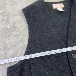 May include: A dark charcoal gray vest with a V-neckline and a single visible button. A white measuring tape is placed across the front, showing a width measurement of approximately 19 inches from the side seam to the center button placket.