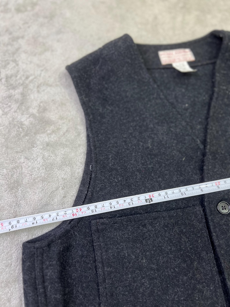 May include: A dark charcoal gray vest with a V-neckline and a single visible button. A white measuring tape is placed across the front, showing a width measurement of approximately 19 inches from the side seam to the center button placket.