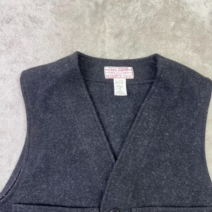 May include: A dark charcoal grey wool vest featuring a V-neckline, a single front button closure, and two large patch pockets. A white rectangular label at the inner neckline reads Filson Garment C.C. Filson Co. Seattle with additional care text.