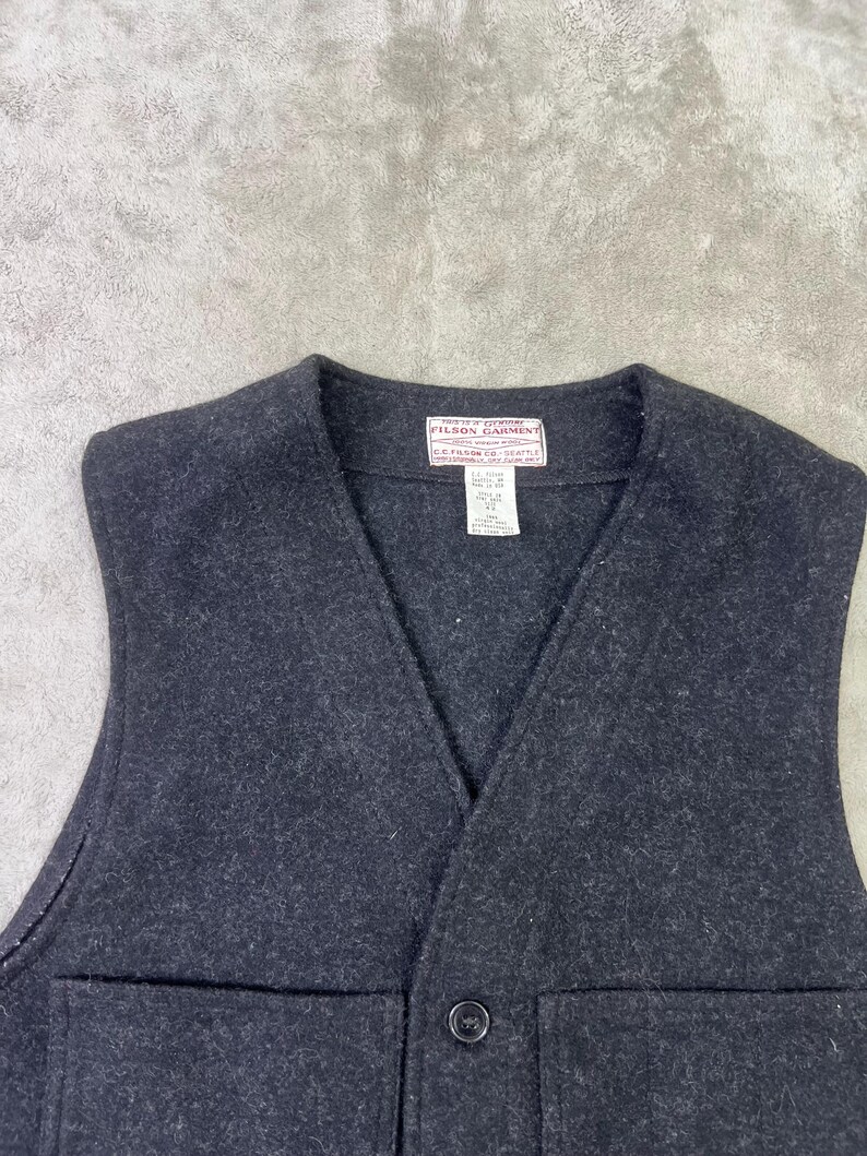 May include: A dark charcoal grey wool vest featuring a V-neckline, a single front button closure, and two large patch pockets. A white rectangular label at the inner neckline reads Filson Garment C.C. Filson Co. Seattle with additional care text.