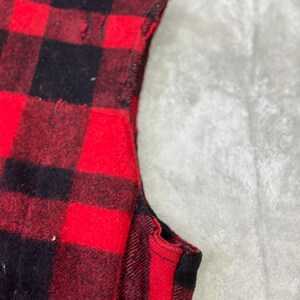 May include: A sleeveless garment featuring a bold red and black buffalo plaid pattern. The fabric displays a classic checkered design with visible stitching along the armhole edge, showcasing the texture and construction of the textile material.