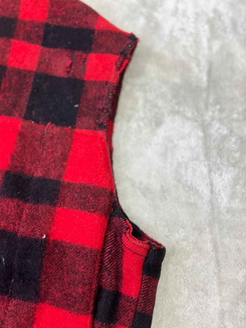 May include: A sleeveless garment featuring a bold red and black buffalo plaid pattern. The fabric displays a classic checkered design with visible stitching along the armhole edge, showcasing the texture and construction of the textile material.