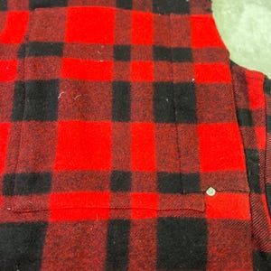 May include: A red and black plaid flannel shirt featuring a chest pocket with a single silver-toned metal rivet detail at the corner. The fabric has a soft, brushed texture with a classic buffalo check pattern throughout the garment.