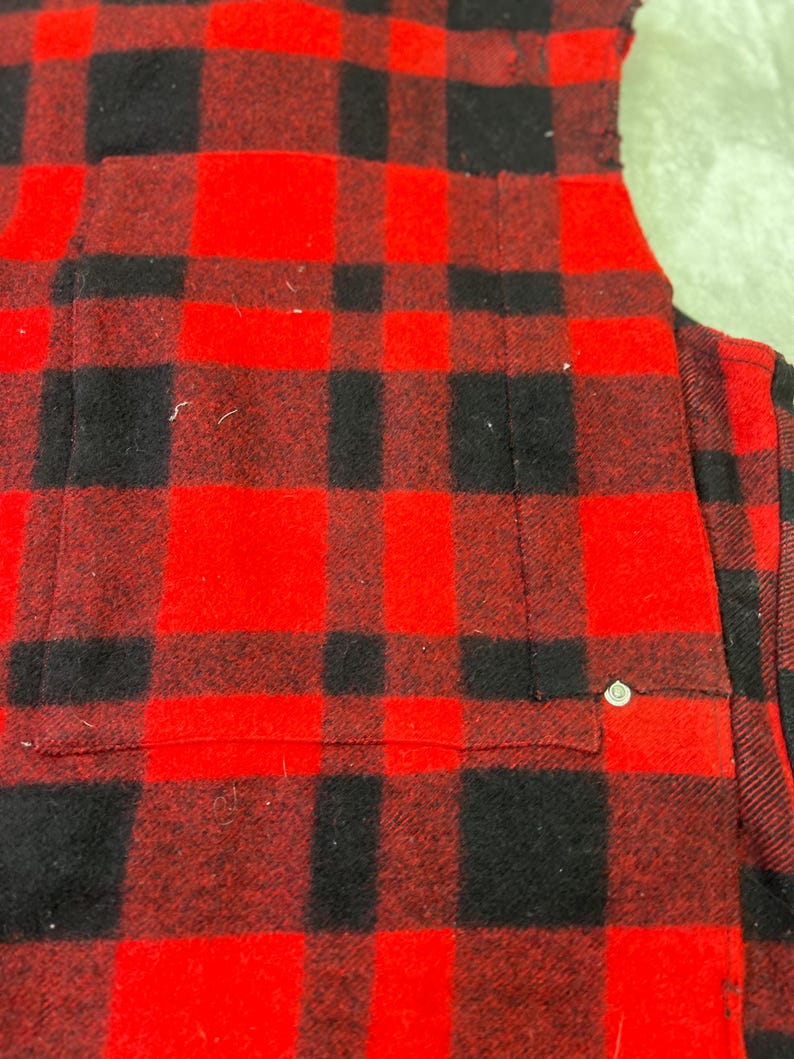 May include: A red and black plaid flannel shirt featuring a chest pocket with a single silver-toned metal rivet detail at the corner. The fabric has a soft, brushed texture with a classic buffalo check pattern throughout the garment.