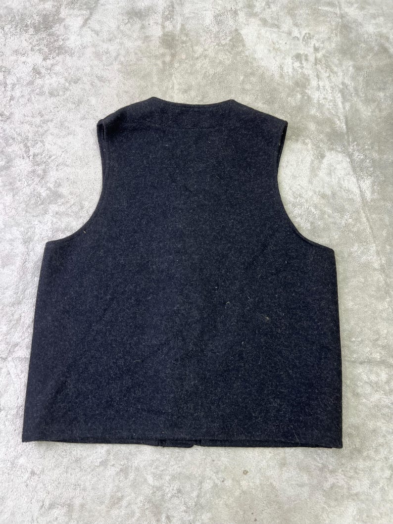 May include: A sleeveless vest in a dark charcoal gray color with a textured, felted appearance. The garment features a rounded neckline, wide arm openings, and a straight hemline, showcasing a simple, solid-colored design without any visible closures.