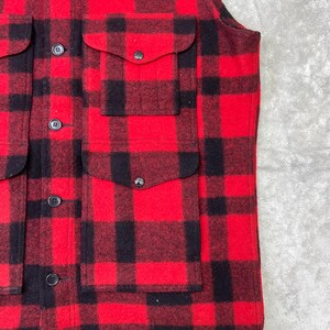 May include: A sleeveless vest featuring a bold red and black buffalo plaid pattern. The garment includes a front button closure with dark buttons and two large patch pockets on the front, each secured with a flap and a matching dark button.