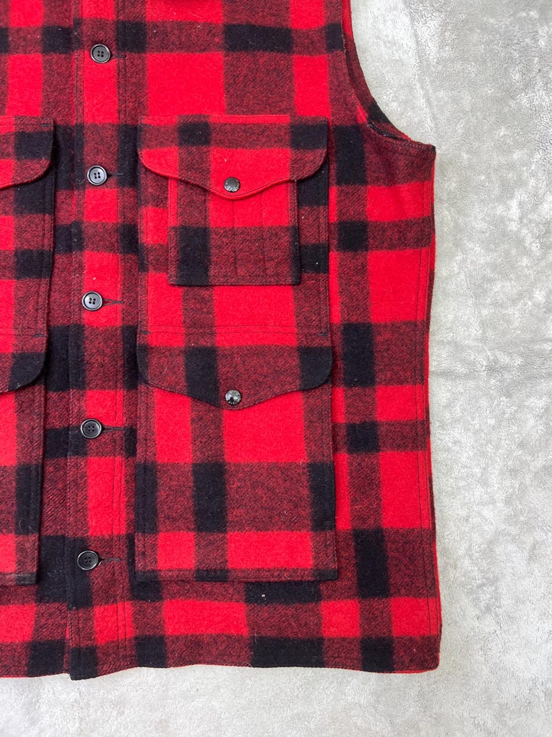 May include: A sleeveless vest featuring a bold red and black buffalo plaid pattern. The garment includes a front button closure with dark buttons and two large patch pockets on the front, each secured with a flap and a matching dark button.