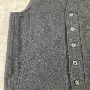 May include: A dark charcoal gray vest made of a textured, felted wool-like material. The garment features a vertical row of five matching dark buttons along the front placket and a large, integrated patch pocket on the side panel.