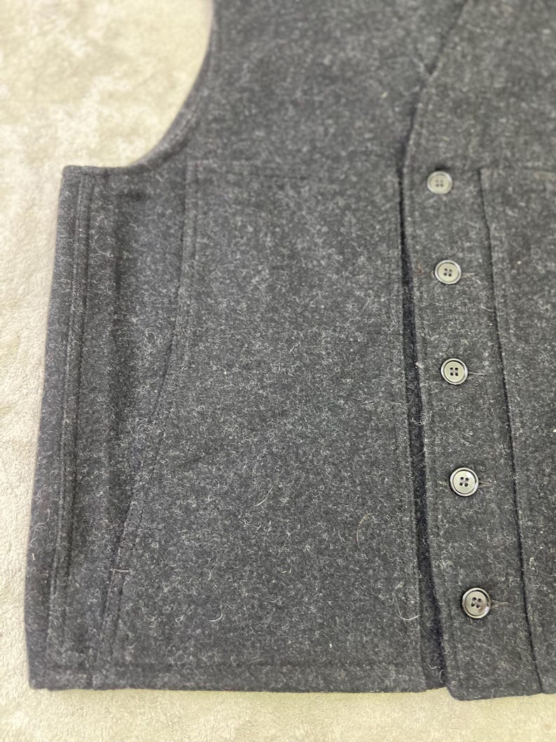 May include: A dark charcoal gray vest made of a textured, felted wool-like material. The garment features a vertical row of five matching dark buttons along the front placket and a large, integrated patch pocket on the side panel.