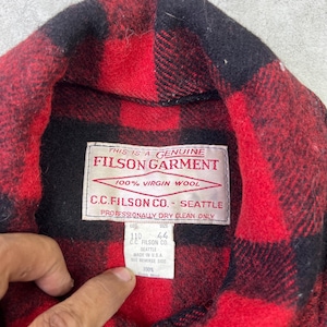 May include: A red and black buffalo plaid wool shirt featuring a white interior label that reads This is a Genuine Filson Garment, 100% Virgin Wool, C.C. Filson Co. Seattle, Professionally Dry Clean Only, with a secondary tag indicating size 44.