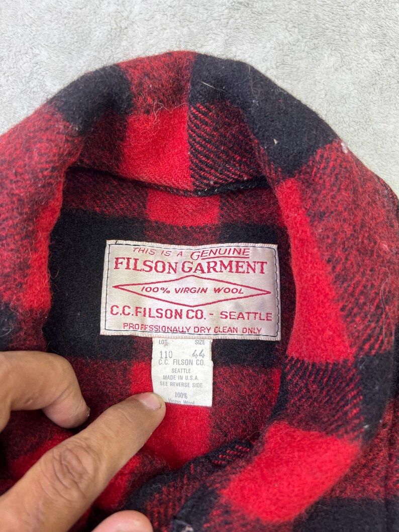 May include: A red and black buffalo plaid wool shirt featuring a white interior label that reads This is a Genuine Filson Garment, 100% Virgin Wool, C.C. Filson Co. Seattle, Professionally Dry Clean Only, with a secondary tag indicating size 44.