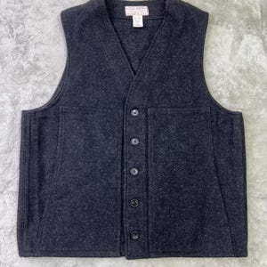 May include: A dark charcoal gray wool vest featuring a V-neckline, a five-button front closure, and two large rectangular patch pockets on the front. A white rectangular brand label with black text is sewn inside the center of the neckline.