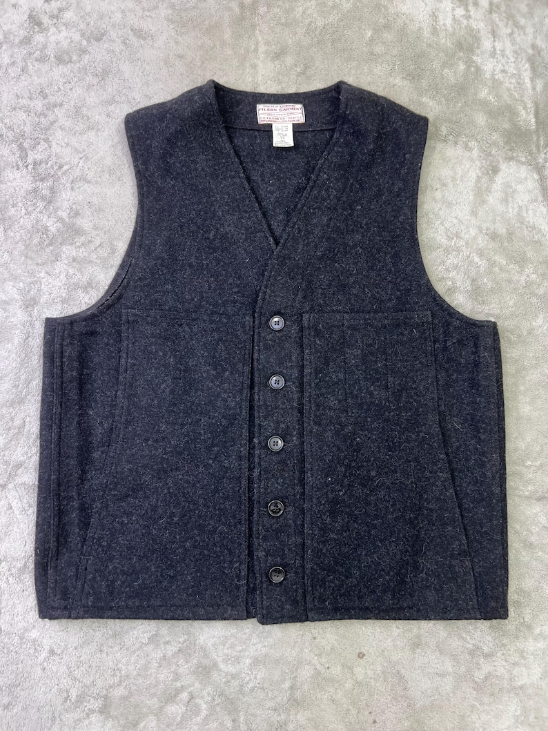 May include: A dark charcoal gray wool vest featuring a V-neckline, a five-button front closure, and two large rectangular patch pockets on the front. A white rectangular brand label with black text is sewn inside the center of the neckline.