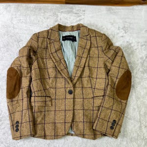 Puede incluir: Una blazer marrón a cuadros con coderas de ante. La blazer tiene un cierre de un solo botón y tres botones en cada puño. El forro interior es de color azul claro. La etiqueta de la marca dice "ZARA BASIC".