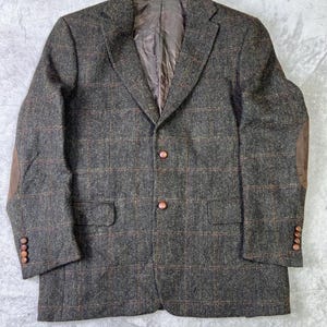 Op de afbeelding: Een tweed blazer in grijstinten, bruin en rood met een ruitpatroon. De blazer heeft bruine elleboogstukken, een revers en bruine knopen. De binnenvoering is van bruine stof.