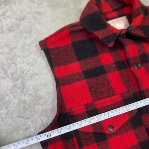 May include: A sleeveless red and black plaid shirt featuring a pointed collar, a front button closure, and a chest pocket with a buttoned flap. A measuring tape is placed across the chest, showing a width measurement of approximately 24 inches.