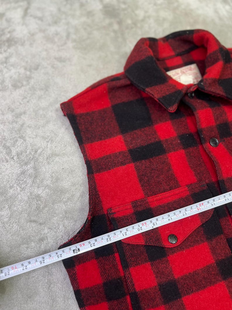 May include: A sleeveless red and black plaid shirt featuring a pointed collar, a front button closure, and a chest pocket with a buttoned flap. A measuring tape is placed across the chest, showing a width measurement of approximately 24 inches.