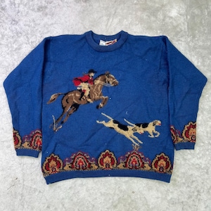 Puede incluir: Jersey de punto azul con un diseño de un jinete a caballo y un perro. Los puños y el dobladillo tienen un patrón decorativo en rojo, marrón y crema. El jersey es de estilo clásico.
