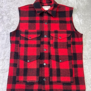 May include: A sleeveless button-up vest featuring a bold red and black buffalo plaid pattern. The garment includes a pointed collar, a central button placket, and four large front pockets with buttoned flaps. A label reads Filson Garment.