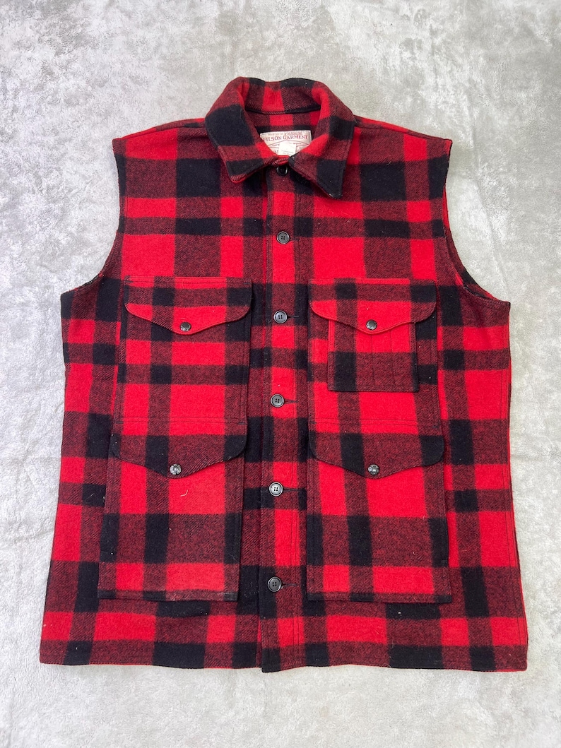 May include: A sleeveless button-up vest featuring a bold red and black buffalo plaid pattern. The garment includes a pointed collar, a central button placket, and four large front pockets with buttoned flaps. A label reads Filson Garment.