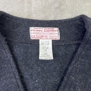 May include: A dark charcoal gray vest made of 100% virgin wool. The garment features a white interior label with red text reading THIS IS A GENUINE FILSON GARMENT 100% VIRGIN WOOL C.C. FILSON CO. SEATTLE PROFESSIONALLY DRY CLEAN ONLY and a size 42 tag.