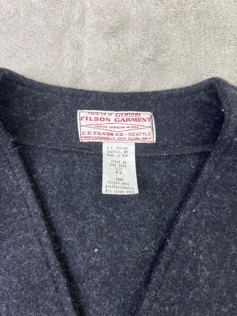 May include: A dark charcoal gray vest made of 100% virgin wool. The garment features a white interior label with red text reading THIS IS A GENUINE FILSON GARMENT 100% VIRGIN WOOL C.C. FILSON CO. SEATTLE PROFESSIONALLY DRY CLEAN ONLY and a size 42 tag.