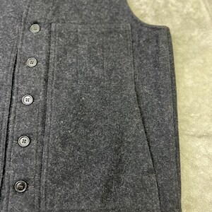 May include: A dark charcoal gray vest made of a thick, textured felted fabric. The garment features a V-neckline, a vertical row of five matching dark buttons down the front, and a large, rectangular patch pocket on the side of the front panel.