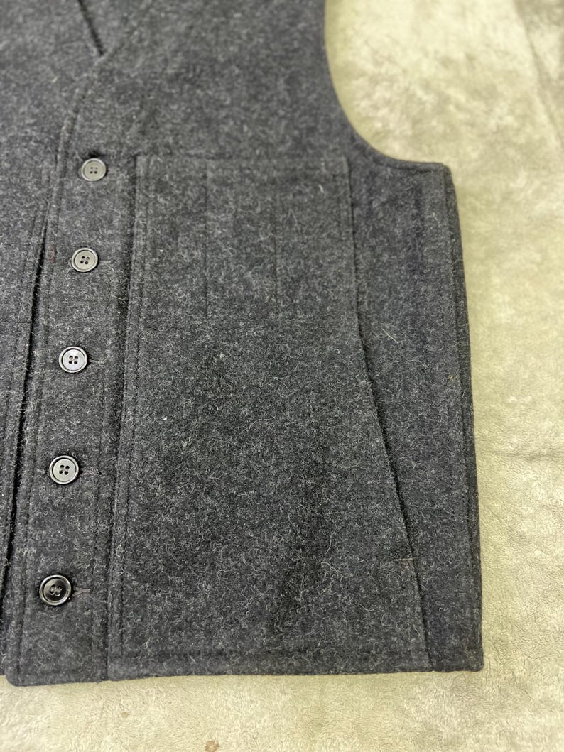 May include: A dark charcoal gray vest made of a thick, textured felted fabric. The garment features a V-neckline, a vertical row of five matching dark buttons down the front, and a large, rectangular patch pocket on the side of the front panel.