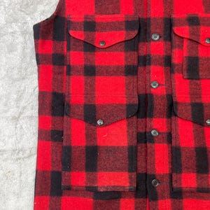 May include: A sleeveless vest featuring a bold red and black buffalo plaid pattern. The garment includes two large front pockets with button-down flaps and a central vertical row of dark buttons for closure, constructed from a textured fabric.