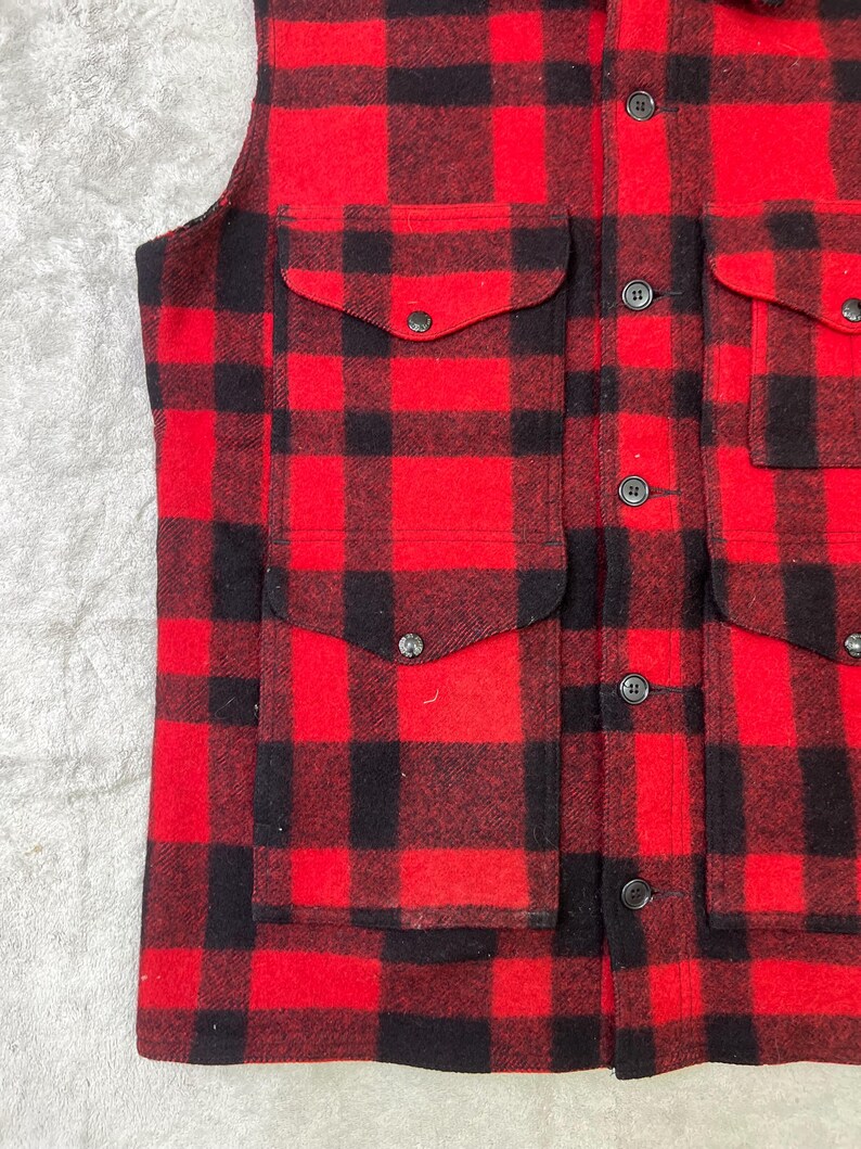 May include: A sleeveless vest featuring a bold red and black buffalo plaid pattern. The garment includes two large front pockets with button-down flaps and a central vertical row of dark buttons for closure, constructed from a textured fabric.